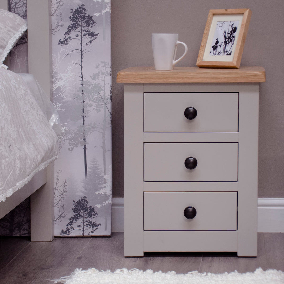 Grey bedside table with three drawers, wooden top, cup and photo frame on bedroom floor