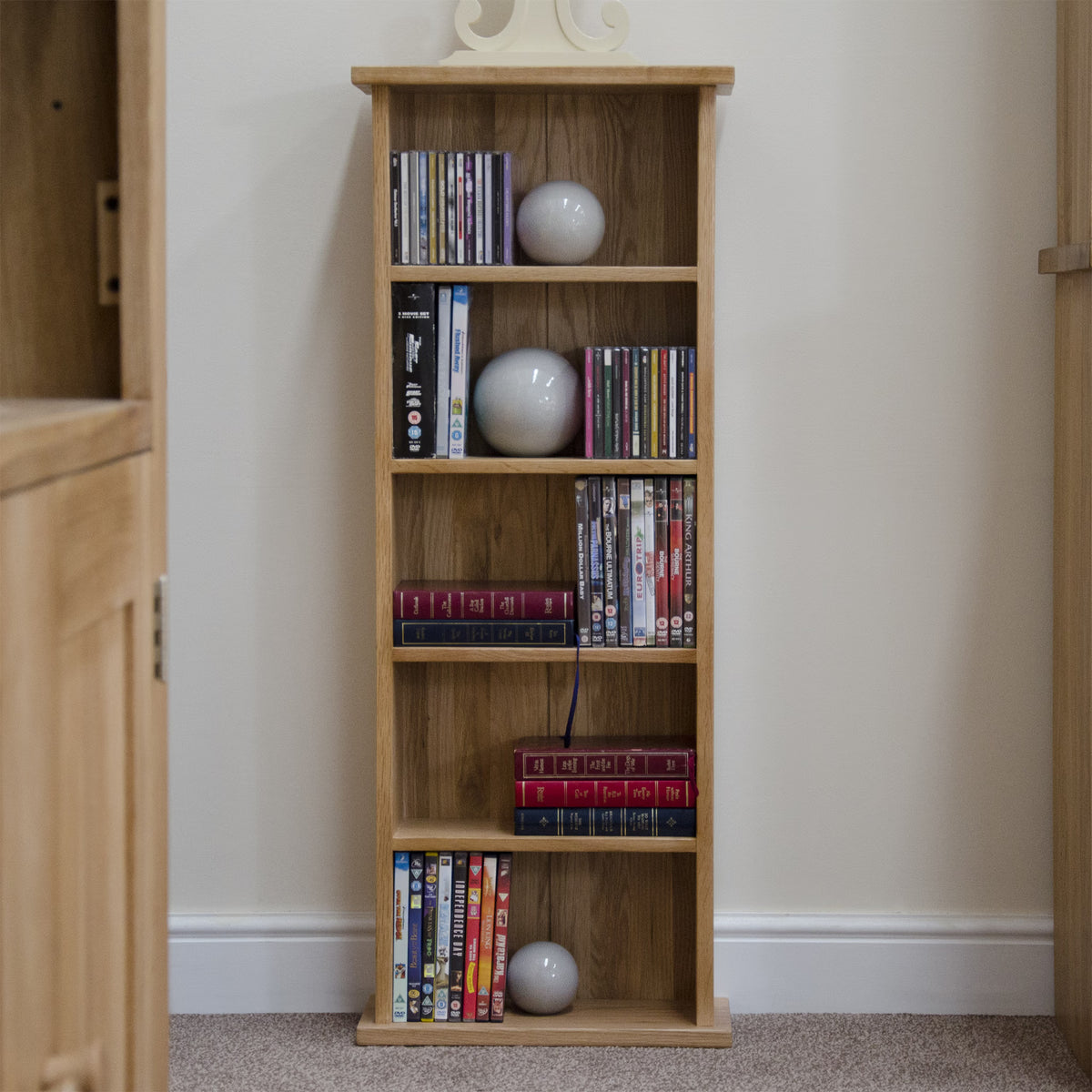 Slim oak bookcase with shelves for DVDs, books, decor orb, against cream wall on carpet