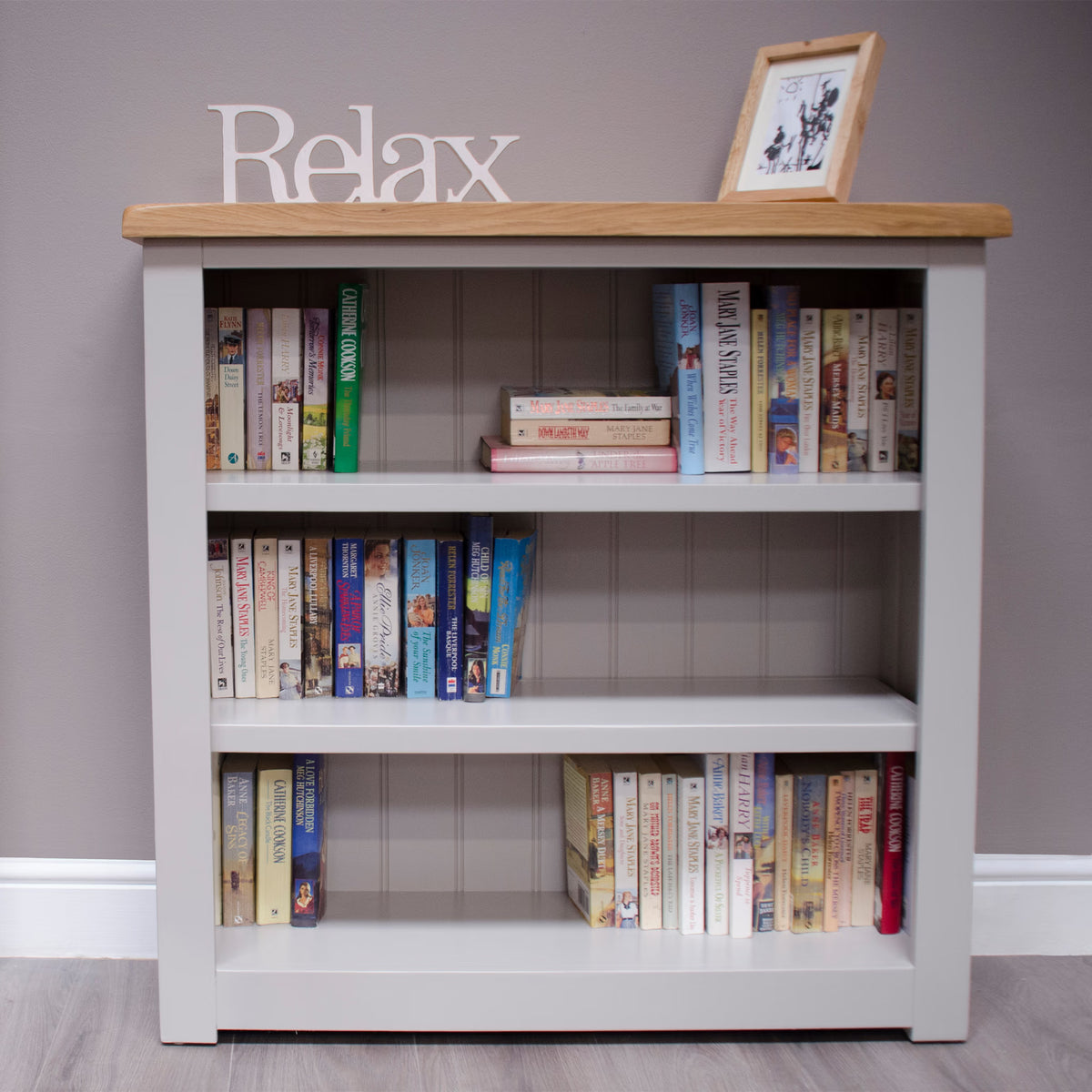 Grey wooden bookcase with oak top, three shelves filled with books, wall mounted decor