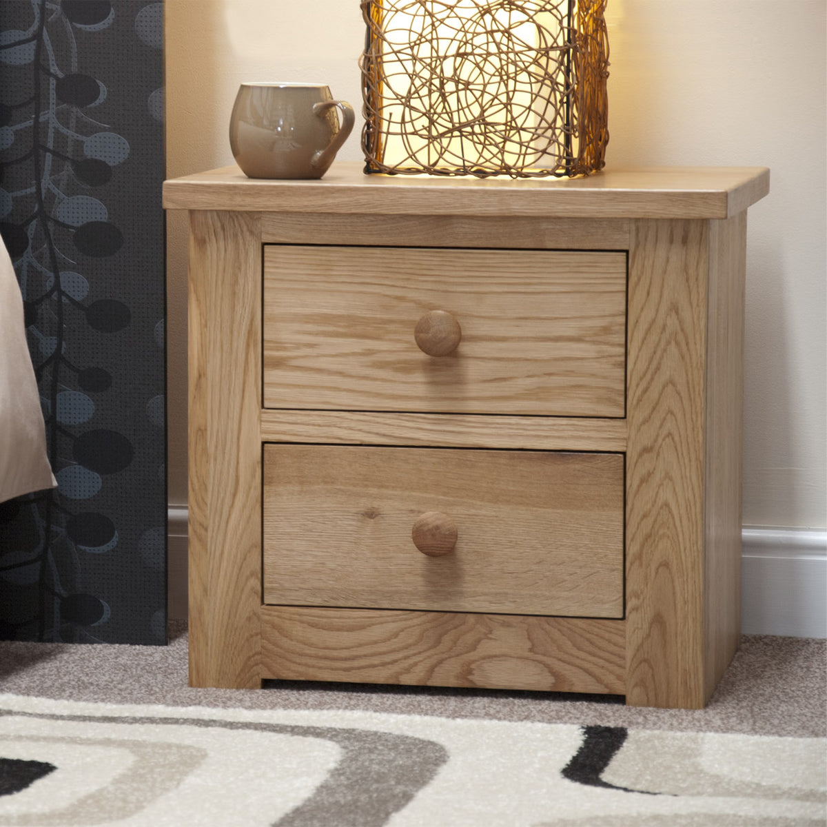 Oak bedside table with two drawers, lamp and mug on top, next to patterned curtain