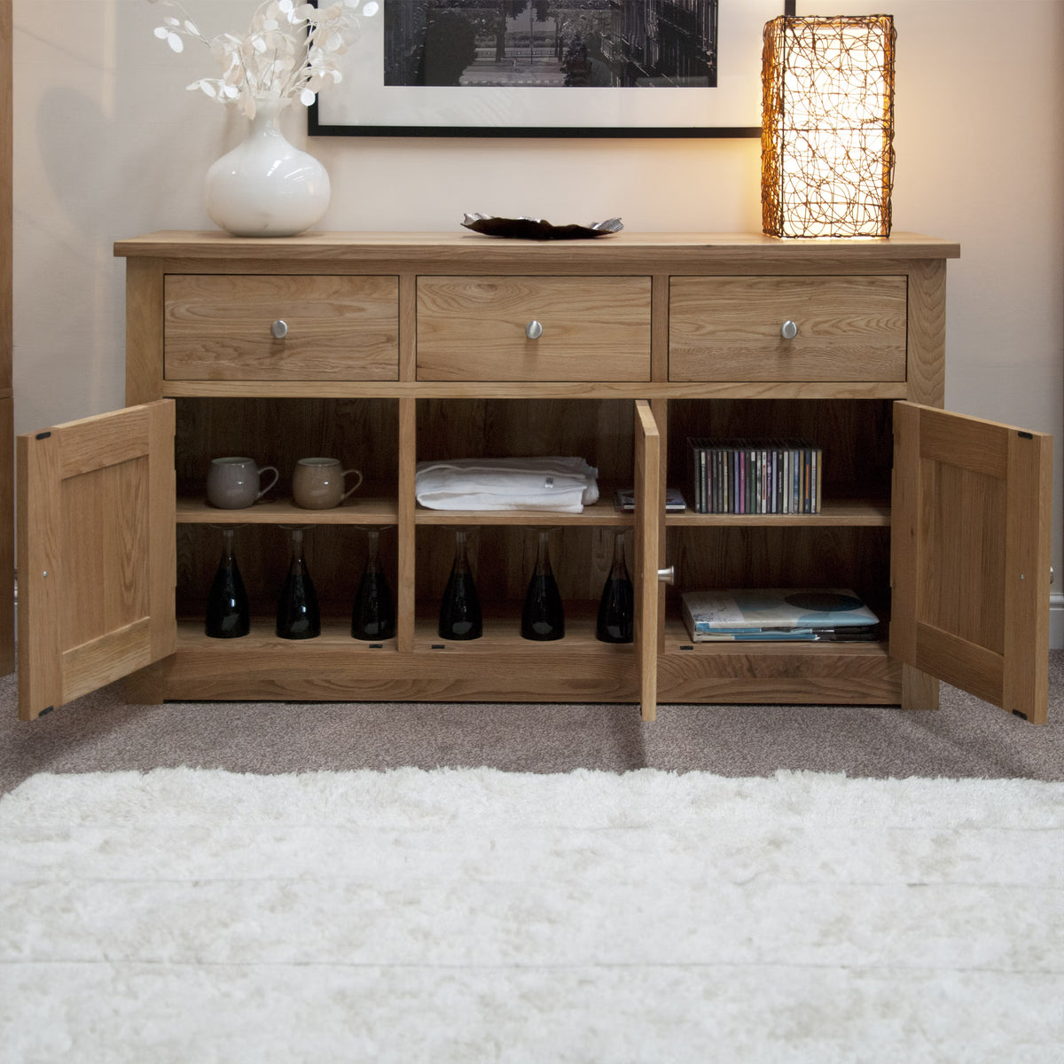 Oak sideboard with three drawers, open cupboard doors, mugs, bottles, and lamp on top