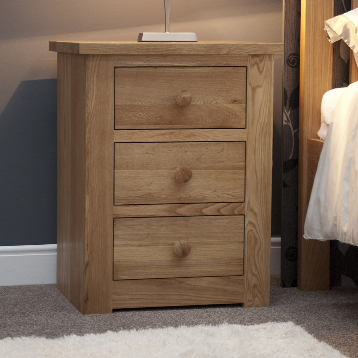 Oak bedside table with three drawers and round wooden handles in bedroom setting