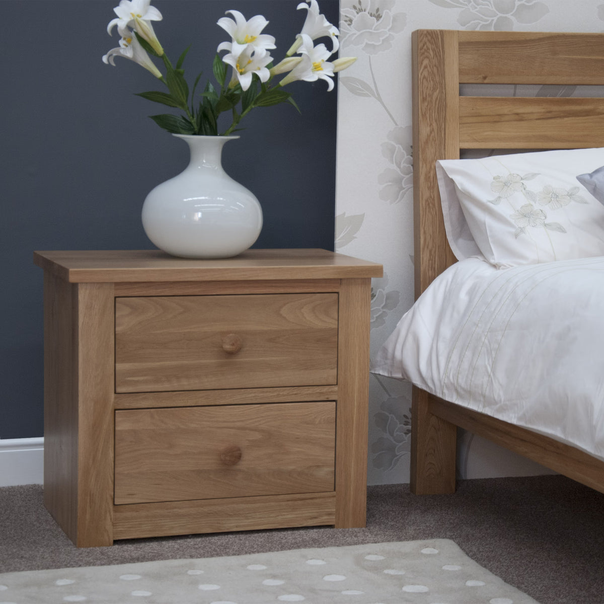 Oak bedside table with two drawers beside bed, white vase with lilies on top