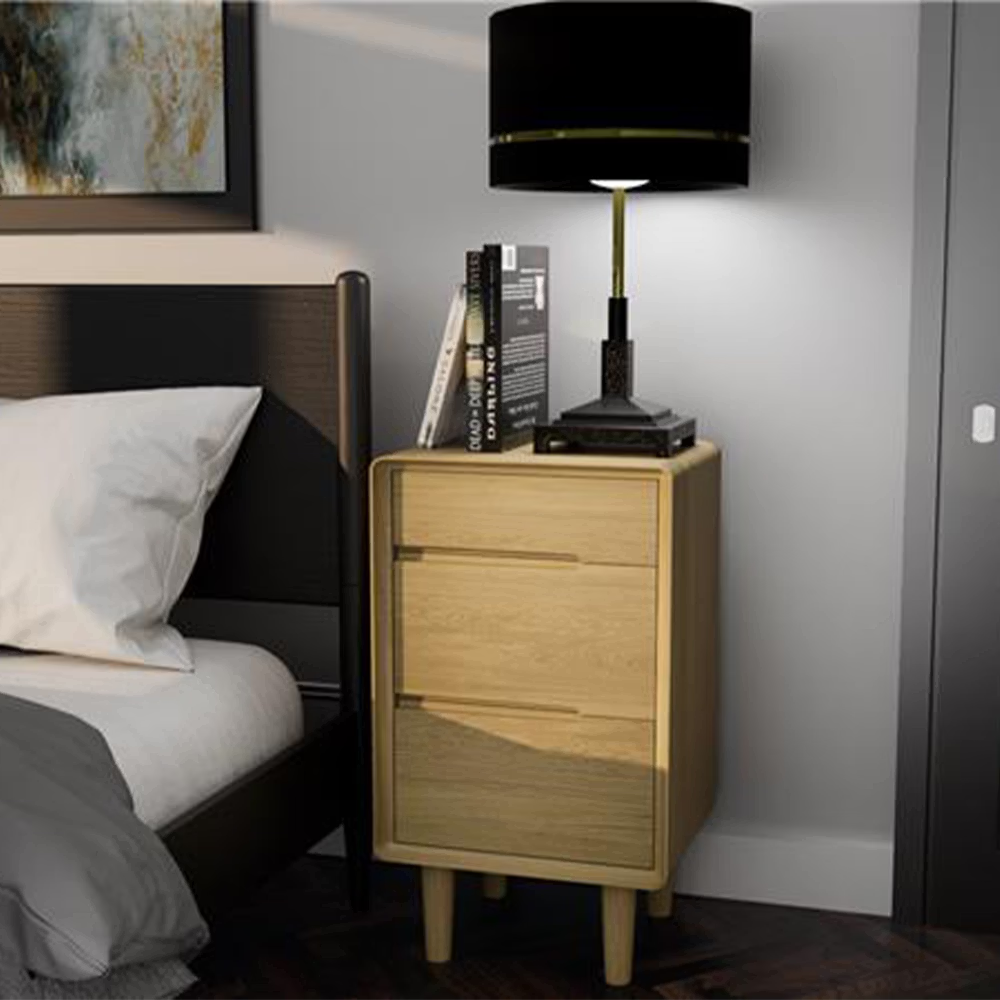Oak bedside table with three drawers, black lamp and books in modern bedroom