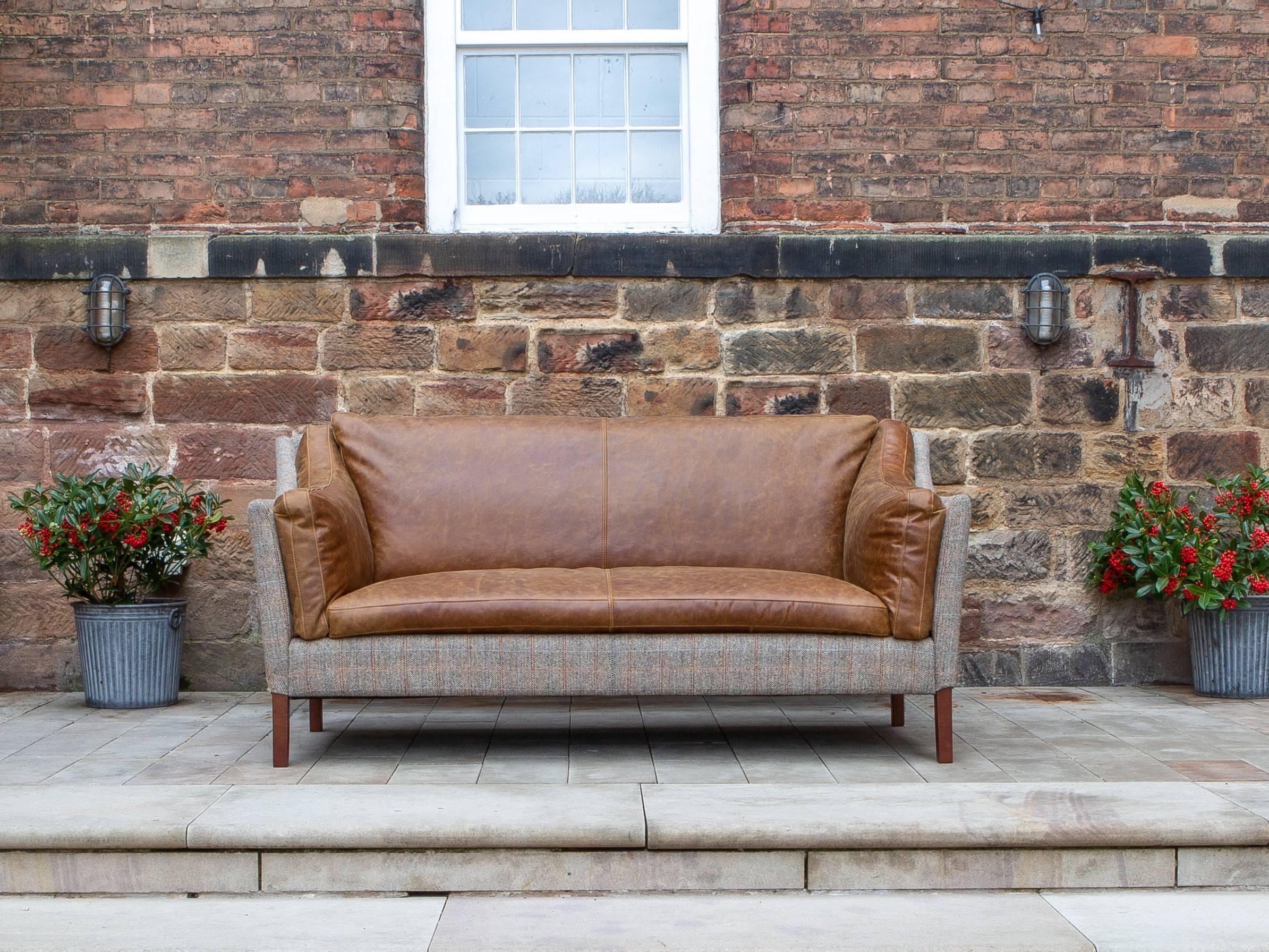 Tan leather and fabric sofa with wooden legs on stone patio against brick wall