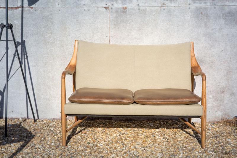 Beige two-seater sofa with wooden frame and brown leather seat cushions on gravel