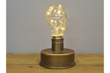 Industrial light with a round bronze metal base and a clear bulb containing warm white string lights on a wooden surface.