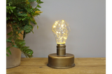 Metal base industrial-style table light with clear bulb and warm white string lights, shown from the front on wooden surface.