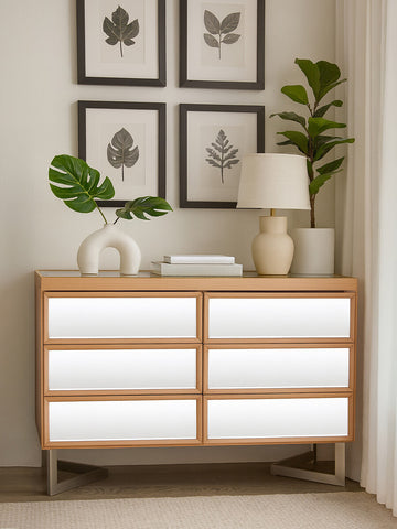 A stylish six-drawer chest with a light wood finish, featuring smooth white drawer fronts and sleek metal handles. The chest is positioned against a wall adorned with framed botanical prints. A decorative lamp and a circular white sculpture sit on top of the chest, alongside a green potted plant, creating a modern and inviting atmosphere