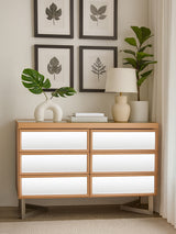 A stylish six-drawer chest with a light wood finish, featuring smooth white drawer fronts and sleek metal handles. The chest is positioned against a wall adorned with framed botanical prints. A decorative lamp and a circular white sculpture sit on top of the chest, alongside a green potted plant, creating a modern and inviting atmosphere