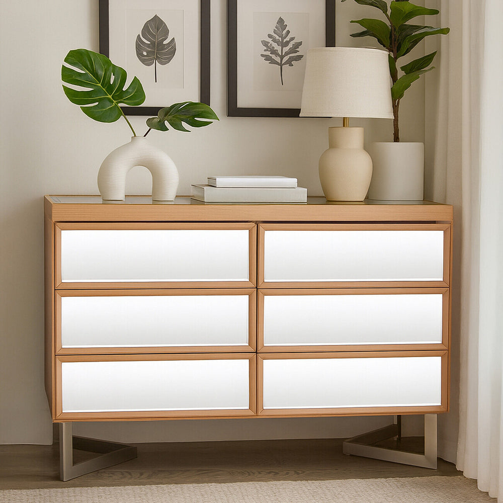 A modern six-drawer chest with a light wood frame and white drawer fronts, positioned against a wall. The chest features sleek lines and a minimalist design, complemented by a decorative lamp, a small plant, and a stack of books on top. Two framed artworks are displayed on the wall above the chest
