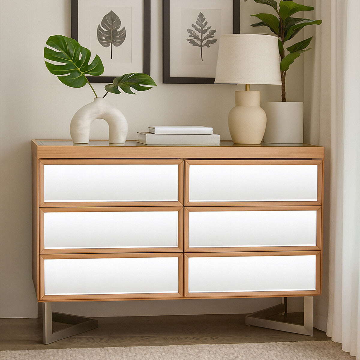 A modern six-drawer chest with a light wood frame and white drawer fronts, positioned against a wall. The chest features sleek lines and a minimalist design, complemented by a decorative lamp, a small plant, and a stack of books on top. Two framed artworks are displayed on the wall above the chest