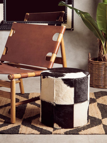 A black and white hair on leather stool with a geometric pattern sits next to a wooden chair on a patterned rug. The stool features a cylindrical shape and is made from leather, showcasing a mix of black and white patches. A green plant is visible in the background, adding a touch of nature to the setting