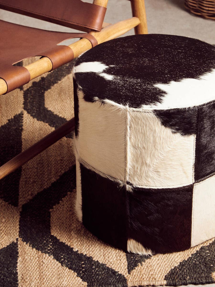 A round stool featuring a black and white hair-on-leather design, with a patchwork pattern of varying sizes. The stool is placed on a geometric patterned rug, alongside a wooden chair with a leather seat