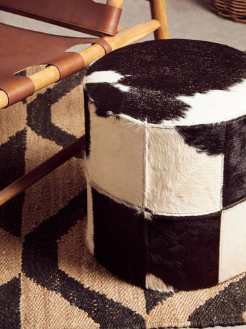 A round stool featuring a black and white hair-on-leather design, with a patchwork pattern of varying sizes. The stool is placed on a geometric patterned rug, alongside a wooden chair with a leather seat