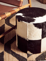 A round stool featuring a black and white hair-on-leather design, with a patchwork pattern of varying sizes. The stool is placed on a geometric patterned rug, alongside a wooden chair with a leather seat