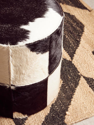 Black and white hair-on leather stool with a cylindrical shape, featuring a patchwork design of varying shades of black and cream. The stool is placed on a textured, geometric patterned rug in neutral tones