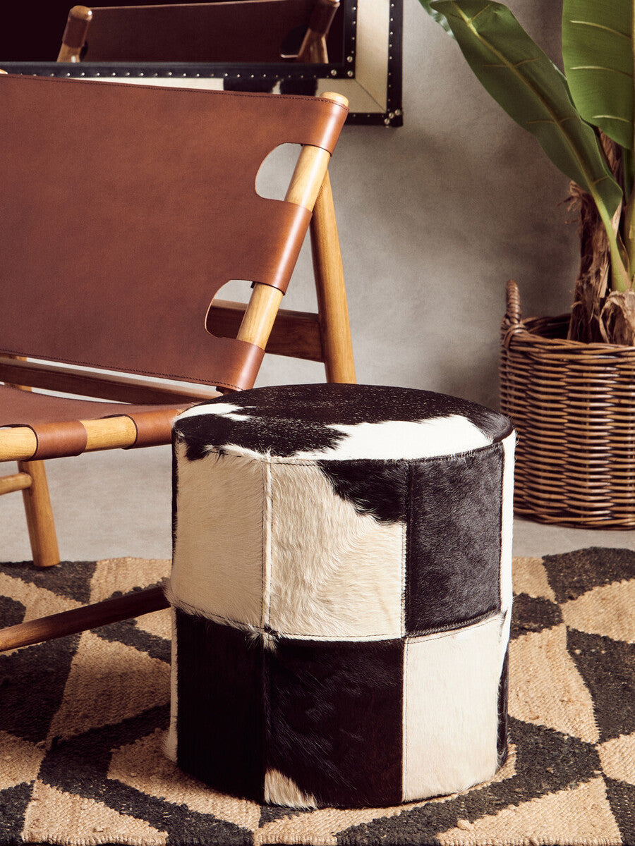 Black and white hair-on leather stool with a patchwork design, featuring a cylindrical shape. The stool is placed on a patterned rug, next to a wooden chair and a potted plant in the background