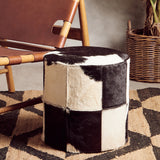 A cylindrical stool featuring a patchwork design of black and white hair-on leather, set on a textured rug. In the background, a wooden chair and a woven basket are partially visible, creating a cozy interior setting