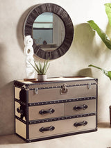Black and white hair-on-leather 3-drawer cabinet with decorative metal accents and handles, positioned against a light-colored wall. A round mirror with a textured frame hangs above the cabinet, and a small potted plant is placed on top alongside a decorative sculpture