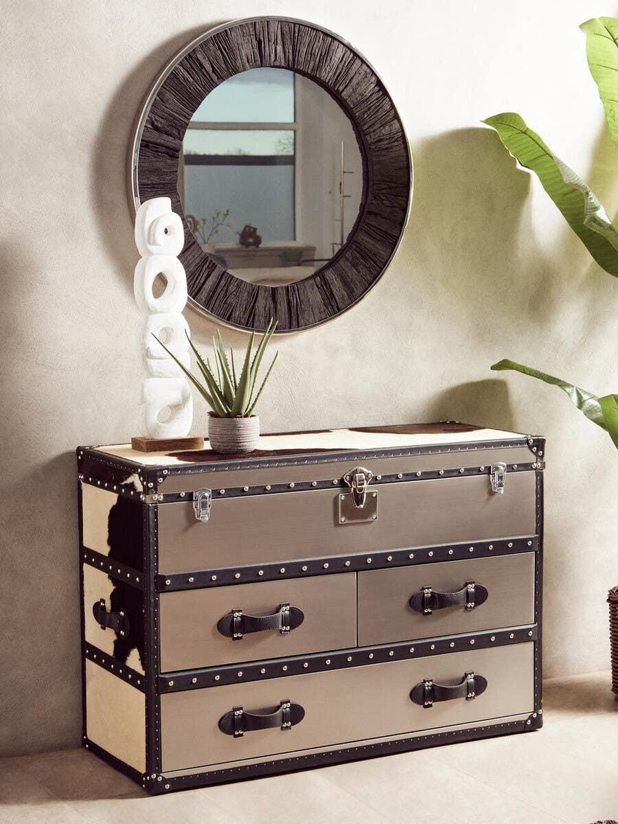 Black and white hair-on-leather 3-drawer cabinet with decorative metal accents and handles, positioned against a light-colored wall. A round mirror with a textured frame hangs above the cabinet, and a small potted plant is placed on top alongside a decorative sculpture
