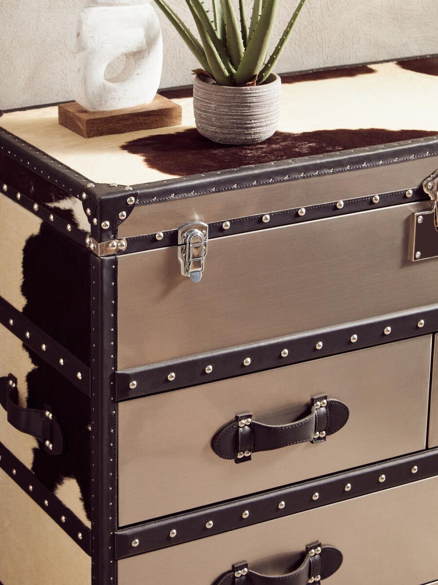 Black and white hair-on-leather cabinet featuring three drawers with decorative metal handles and corner accents. The top surface is styled with a small potted plant and a tissue box, set against a light-colored background