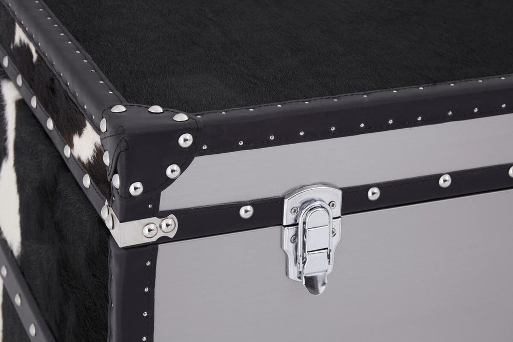 Black and white hair-on-leather cabinet featuring three drawers, with a sleek design and metal accents. The corners are reinforced with silver studs, and a sturdy latch is visible on the front. The surface has a smooth finish, highlighting the contrasting textures of the leather