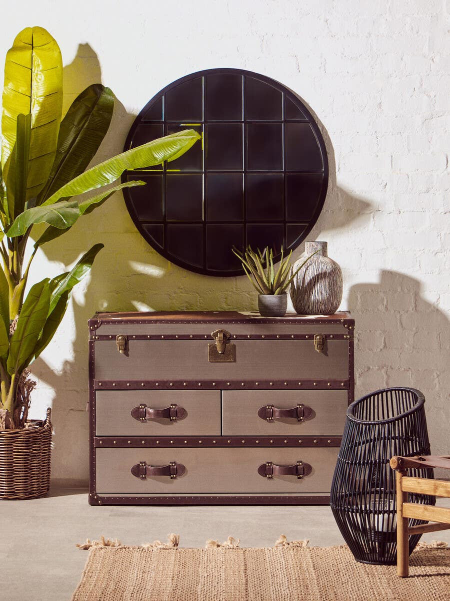 A brown and white cowhide cabinet with three drawers, featuring metal handles, sits against a textured wall. Above the cabinet, a large round black mirror reflects the room. A tall green plant is positioned to the left, and a decorative item is placed on top of the cabinet. A black woven chair is visible to the right, complementing the overall decor