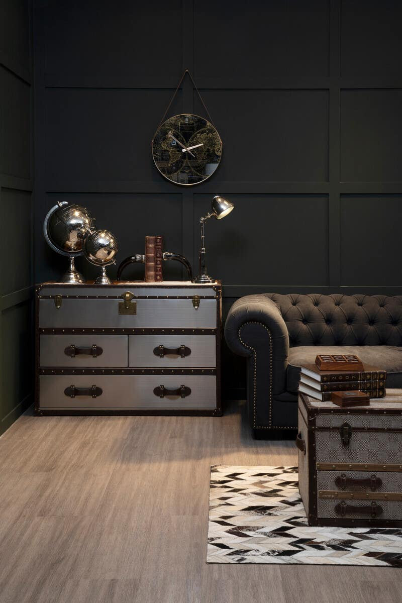 Brown and white cowhide cabinet with two drawers, placed against a dark wall in a stylish living room setting. A black leather chesterfield sofa is visible to the right, and a decorative rug with a geometric pattern is on the floor. A vintage globe and a wooden cross are displayed on top of the cabinet, adding to the room's elegant decor