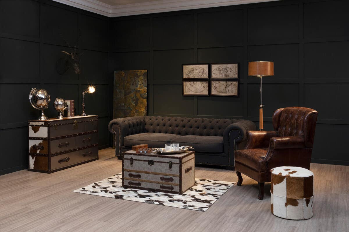 A stylish living room featuring a brown and white cowhide cabinet, a dark gray sofa, a brown leather armchair, and a wooden coffee table. The floor is covered with a light-colored rug, and the walls are painted dark green. Decorative items, including a lamp and framed artwork, are arranged on the cabinet and walls, creating a cozy atmosphere