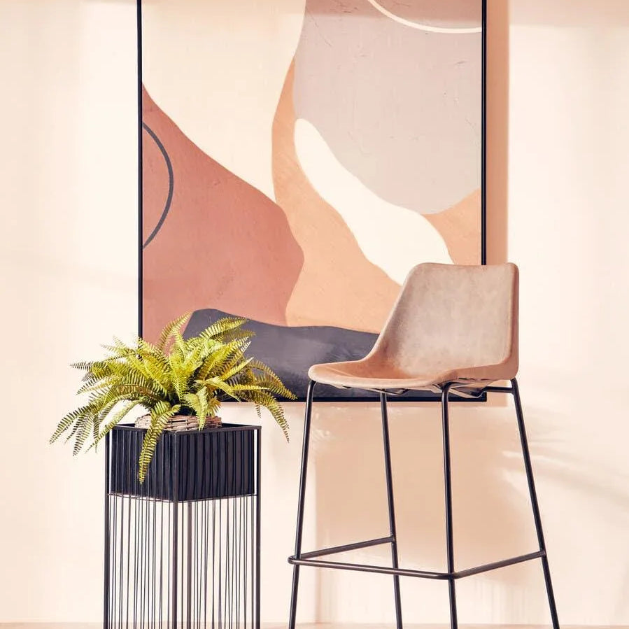 Modern bar stool with beige seat and black legs beside metal plant stand and abstract wall art