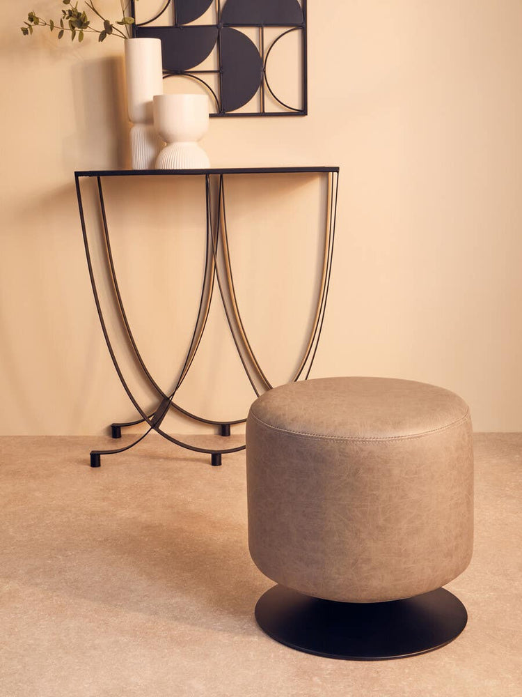 A round pouffe in ash grey faux leather sits on a light-colored floor, next to a modern black metal side table with curved legs. The table holds a white vase and a framed artwork, creating a stylish and contemporary living space