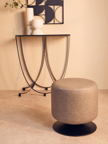 A round pouffe in ash grey faux leather sits on a light-colored floor, next to a modern black metal side table with curved legs. The table holds a white vase and a framed artwork, creating a stylish and contemporary living space