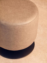 Round pouffe in ash grey faux leather with a smooth surface and a subtle sheen, casting a soft shadow on a light-colored floor