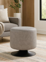Round pouffe in ash grey faux leather with a smooth surface, resting on a black circular base, placed in a cozy living room setting with a light-colored rug and a sofa in the background. Natural light filters through a nearby window, highlighting the pouffe's texture