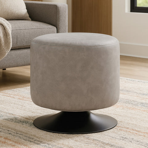 A round pouffe upholstered in ash grey faux leather, featuring a smooth, soft texture and a sturdy black base. The pouffe is placed on a light-colored rug in a cozy living space, with a sofa and decorative elements visible in the background