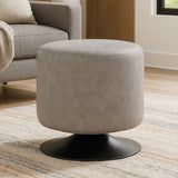 A round pouffe upholstered in ash grey faux leather, featuring a smooth, soft texture and a sturdy black base. The pouffe is placed on a light-colored rug in a cozy living space, with a sofa and decorative elements visible in the background