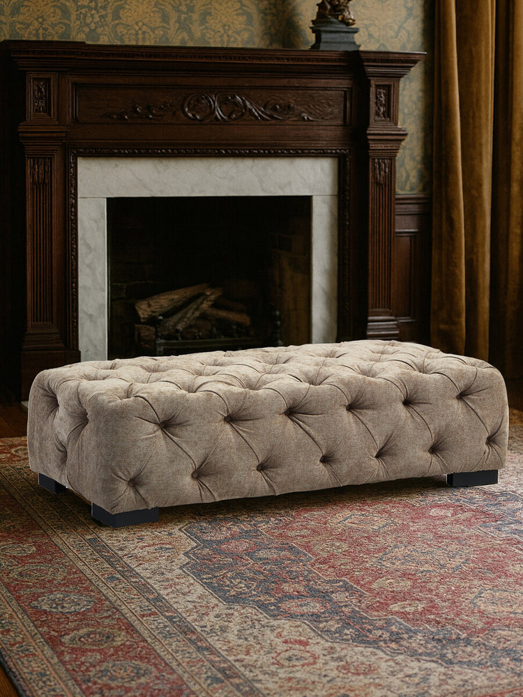 Rectangular footstool upholstered in natural fabric with tufted detailing and decorative buttons, placed in front of a fireplace. The footstool is situated on a patterned area rug, with warm-toned curtains in the background