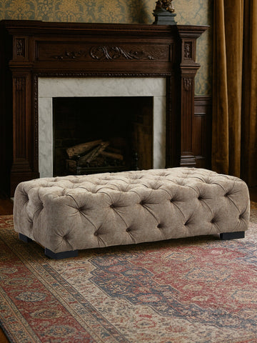 Rectangular footstool upholstered in natural fabric with tufted detailing and decorative buttons, placed in front of a fireplace. The footstool is situated on a patterned area rug, with warm-toned curtains in the background