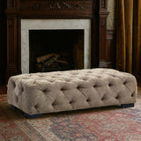 Rectangular footstool upholstered in natural fabric with a tufted design, featuring a soft, cushioned top and dark wooden legs. The footstool is placed in a cozy room setting with a fireplace and patterned rug in the background
