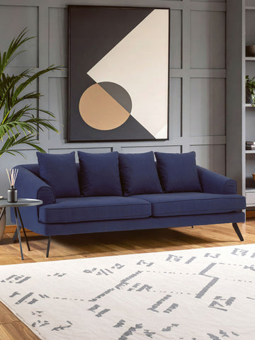 Navy fabric three-seater sofa with plush cushions, sleek metal legs, and a modern design, placed in a stylish living room setting featuring a geometric wall art piece and a patterned area rug. A green plant adds a touch of nature to the decor