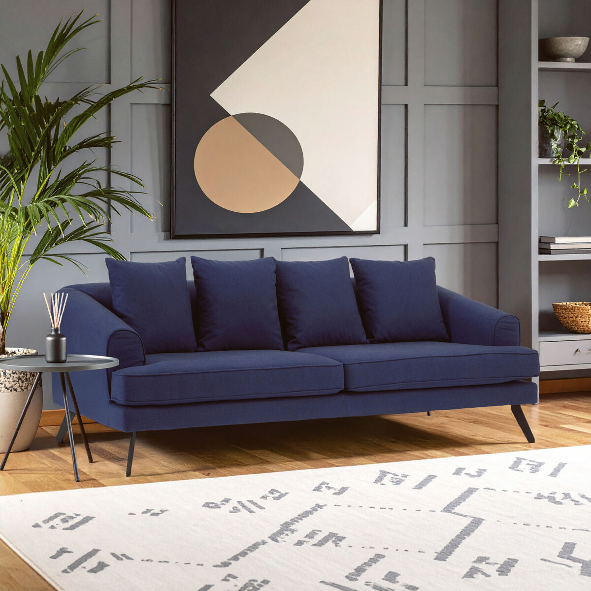 Navy fabric three-seater sofa with four plush cushions, set against a modern interior featuring a geometric wall art piece and a potted plant, placed on a patterned area rug