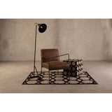 A stylish grey leather patchwork pouffe placed on a geometric patterned rug, accompanied by a modern brown armchair and a sleek floor lamp with a black shade, all set against a textured neutral wall