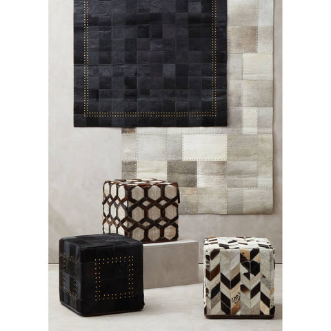 A grey leather patchwork pouffe with a textured surface, featuring a mix of geometric patterns in black and cream. The pouffe is placed on a light-colored floor, surrounded by other decorative pouffes with contrasting designs, against a neutral wall backdrop