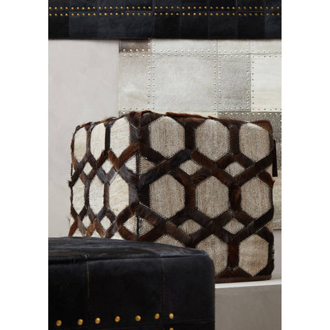 A grey leather patchwork pouffe featuring a geometric hexagonal pattern in varying shades of grey and brown. The pouffe has a soft texture and is set against a neutral background, with additional dark furniture elements partially visible in the foreground