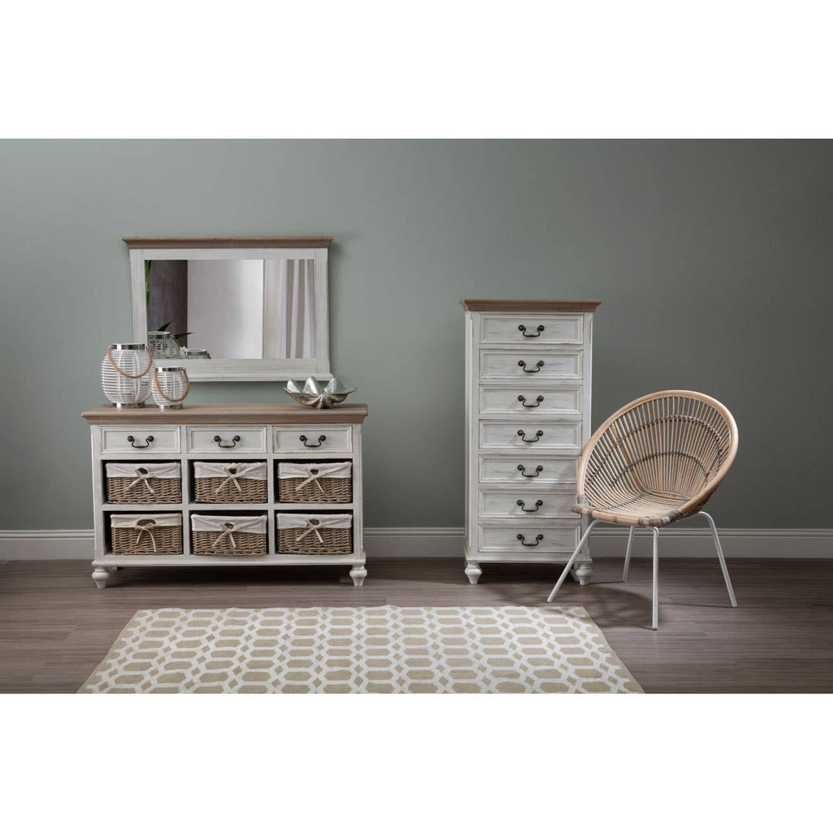 Antique style chest of seven drawers in a light wood finish with a contrasting top, placed against a muted green wall. A matching mirror is positioned above the chest, and a tall, narrow cabinet with similar design is visible beside it. A round, woven chair sits nearby on a light-colored rug