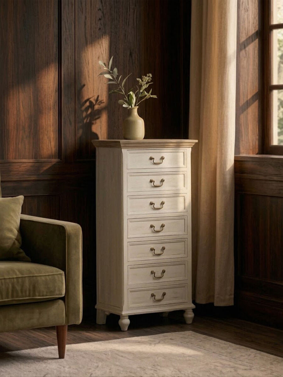 Antique style chest of seven drawers in a light finish, featuring ornate drawer pulls and a smooth top surface, positioned next to a green upholstered armchair in a warmly lit room with wooden walls and large windows. A decorative vase with branches sits atop the chest
