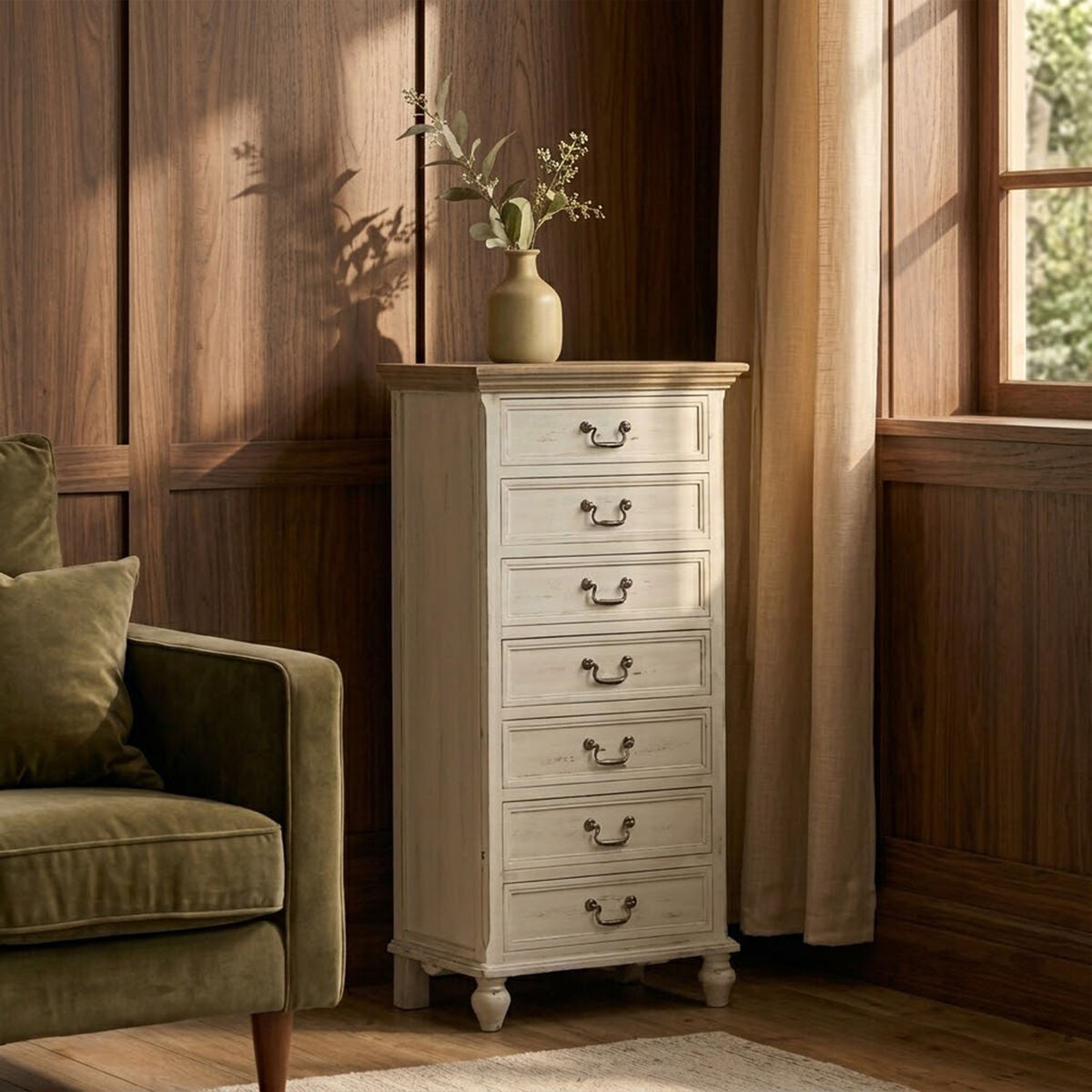 Antique-style chest of seven drawers in a light finish, featuring decorative handles and a smooth top surface. The chest is positioned beside a green upholstered armchair in a warmly lit room with wooden walls and large windows, allowing natural light to illuminate the space. A vase with greenery sits atop the chest