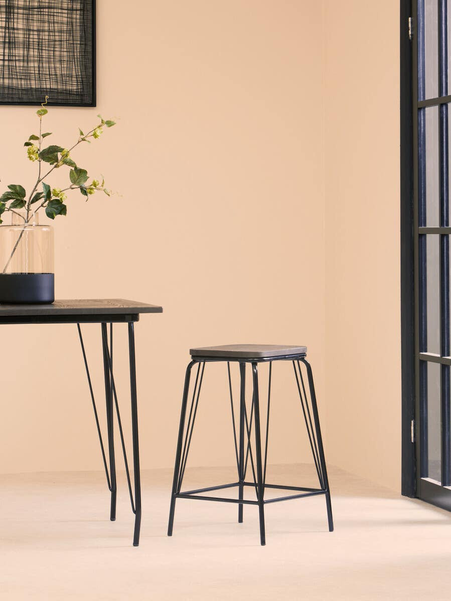 A black metal stool with a simple design and a natural elm wood seat, positioned next to a sleek black table in a softly lit room with a light-colored wall and a large window. A potted plant sits on the table, adding a touch of greenery to the scene