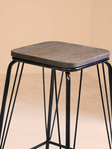 A black metal stool featuring a square elm wood seat, supported by slender, intersecting metal legs, set against a soft, neutral background